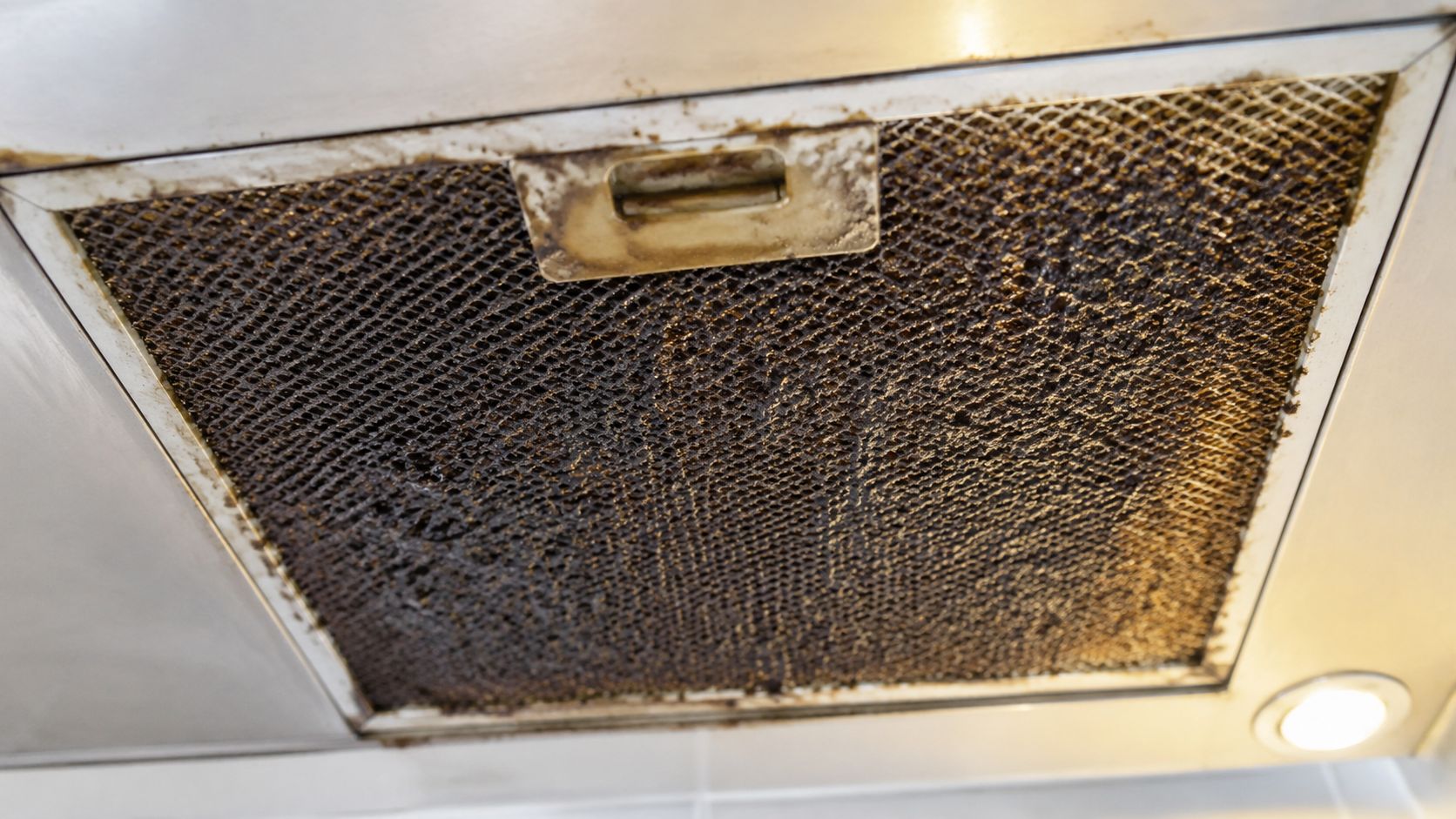 A close-up view of a dirty, grease-covered metal mesh filter inside a kitchen range hood vent.